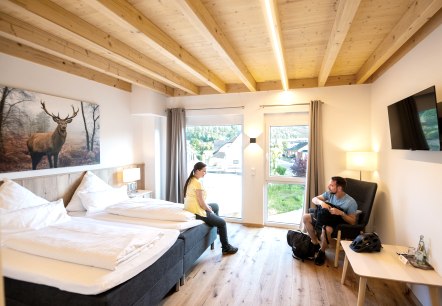 Übernachten Eifel , © Eifeltourismus GmbH,Domink Ketz Hotelzimmer , rechts sitzt ein Mann auf einem Sessel an der Wand ein Fernseher, links sitzt eine Frau auf dem Doppelbett