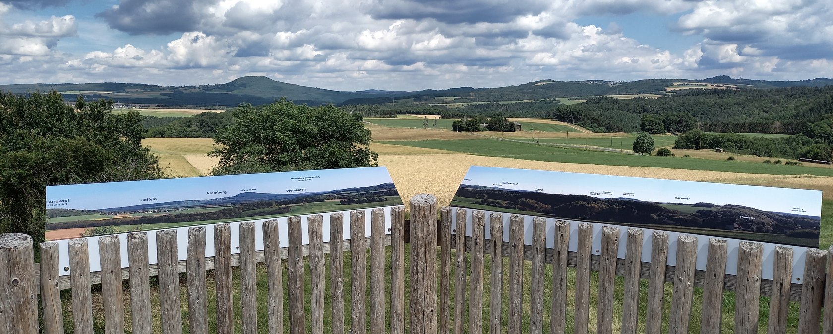 Aussichtsplattform mit Holzzaun, Informationstafeln und Blick auf eine hügelige Landschaft unter blauem Himmel mit Wolken., © Siegfried Müller Aussichtsplattform mit Holzzaun, Informationstafeln und Blick auf eine hügelige Landschaft unter blauem Himmel mit Wolken., © Siegfried Müller