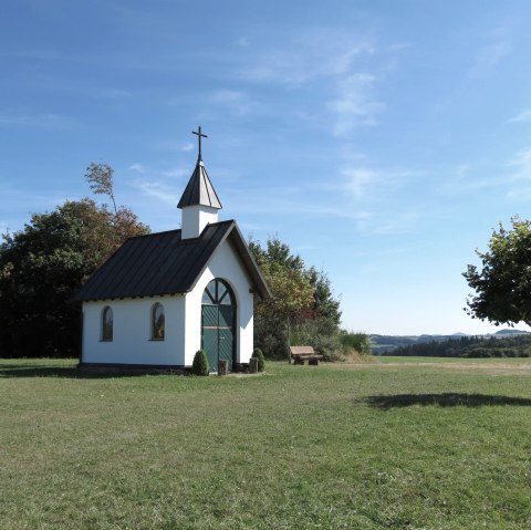 Die Kottenborner Kapelle bei Wershofen steht malerisch auf einer grünen Wiese unter klarem, blauem Himmel. Ein Baum und Bänke umgeben die Kapelle., © VGVAdenau Die Kottenborner Kapelle bei Wershofen steht malerisch auf einer grünen Wiese unter klarem, blauem Himmel. Ein Baum und Bänke umgeben die Kapelle., © VGVAdenau
