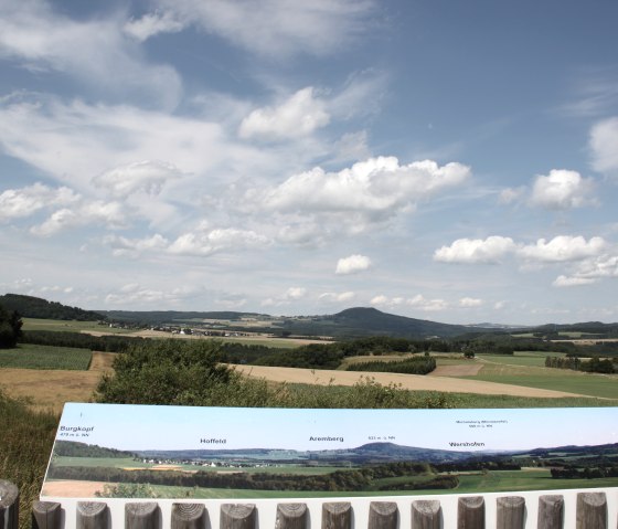 Aussichtsplattform mit Blick auf Felder und Hügel unter blauem Himmel. Ein Schild zeigt Informationen zu sichtbaren Orten., © Uschi Regh Aussichtsplattform mit Blick auf Felder und Hügel unter blauem Himmel. Ein Schild zeigt Informationen zu sichtbaren Orten., © Uschi Regh