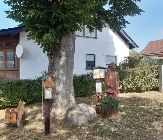 Ein Rastplatz mit einer Holzfigur, Sitzbank und Infotafeln unter einem großen Baum vor einem weißen Haus., © Tourist-Information Hocheifel-Nürburgring,H.Bauerfeind Ein Rastplatz mit einer Holzfigur, Sitzbank und Infotafeln unter einem großen Baum vor einem weißen Haus., © Tourist-Information Hocheifel-Nürburgring,H.Bauerfeind