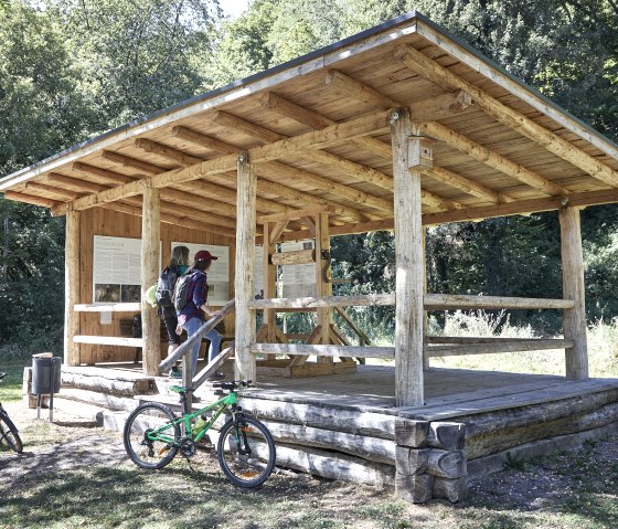 Two people read information boards in a wooden shelter in the countryside. Bicycles are parked next to it, surrounded by trees., © TI Hocheifel-Nürburgring_Jonathan_Andrews Two people read information boards in a wooden shelter in the countryside. Bicycles are parked next to it, surrounded by trees., © TI Hocheifel-Nürburgring_Jonathan_Andrews