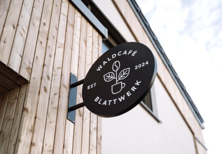 Transparent Eingangsschild Waldcafe`Blattwerk, © Wohllebens Waldakademie rundes Eingangsschild mit dem Namen Waldcafe`Blattwerk Jahreszahl 2024 und einer Tasse aus der eien Blume wächst