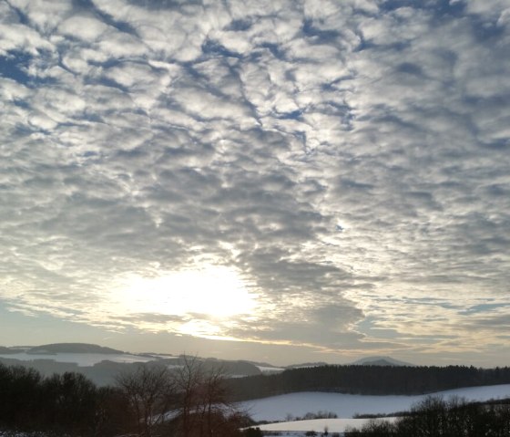 Winterlandschaft in der Eifel mit schneebedeckten Feldern, Bäumen und einem dramatischen, bewölkten Himmel bei Sonnenuntergang., © TI Hocheifel-Nürburgring Winterlandschaft in der Eifel mit schneebedeckten Feldern, Bäumen und einem dramatischen, bewölkten Himmel bei Sonnenuntergang., © TI Hocheifel-Nürburgring