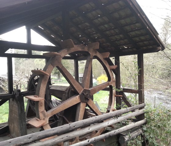 Une vieille roue à eau sous un toit en bois, entourée d'un paysage verdoyant et d'une rivière en arrière-plan., © TI Hocheifel-Nürburgring©J.Hecken Une vieille roue à eau sous un toit en bois, entourée d'un paysage verdoyant et d'une rivière en arrière-plan., © TI Hocheifel-Nürburgring©J.Hecken