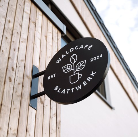 Transparent Eingangsschild Waldcafe`Blattwerk, © Wohllebens Waldakademie rundes Eingangsschild mit dem Namen Waldcafe`Blattwerk Jahreszahl 2024 und einer Tasse aus der eien Blume wächst