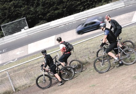Vier mountainbikers staan bij een hek en kijken naar een auto die voorbij rijdt op een racecircuit., © Markus Risse Vier mountainbikers staan bij een hek en kijken naar een auto die voorbij rijdt op een racecircuit., © Markus Risse
