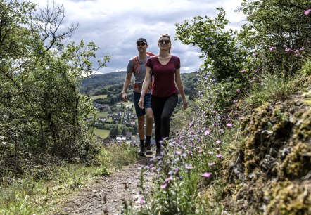 Zwei Wanderer auf einem blumengesäumten Pfad am AhrSteig bei Schuld. Im Hintergrund sind Hügel und ein Dorf zu sehen., © Christopher Pfromm Zwei Wanderer auf einem blumengesäumten Pfad am AhrSteig bei Schuld. Im Hintergrund sind Hügel und ein Dorf zu sehen., © Christopher Pfromm
