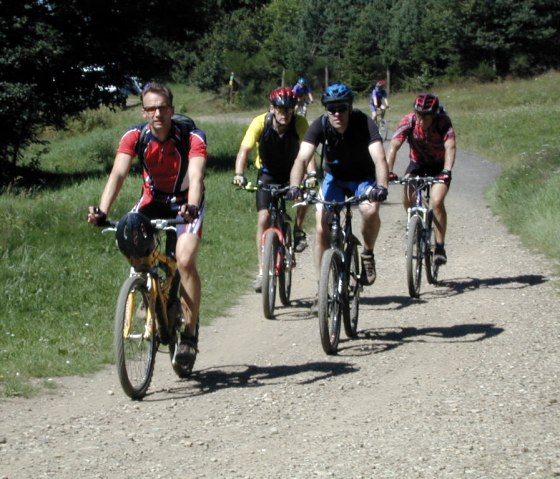 Vijf mountainbikers rijden over een grindpad door een groen landschap. Ze dragen helmen en sportieve kleding., © RC Herschbroich Vijf mountainbikers rijden over een grindpad door een groen landschap. Ze dragen helmen en sportieve kleding., © RC Herschbroich