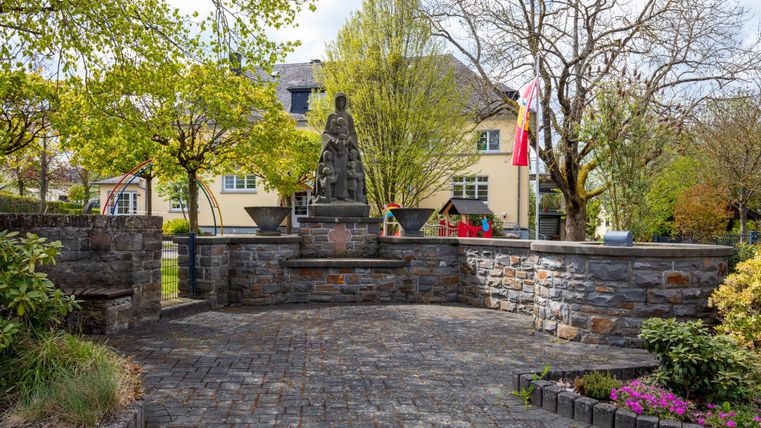 Ein ruhiger Platz mit einer Statue in der Mitte und umgeben von Bäumen und Blumen. Im Hintergrund sind Gebäude und eine grüne Parklandschaft zu sehen.
