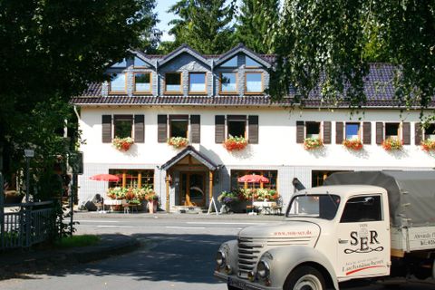 Un charmant restaurant avec des jardinières fleuries et une terrasse accueillante. Au premier plan, un véhicule nostalgique qui contribue à l'ambiance.