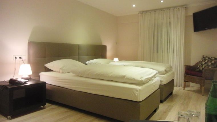 Une chambre d'hôtel moderne avec deux lits jumeaux et un fauteuil confortable. La chambre dispose d'un éclairage chaleureux et de grandes fenêtres avec des rideaux.