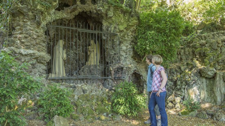 Ein Pärchen steht vor einer steinernen Nische mit einer Gittertüre, die zwei skulpturale Figuren zeigt. Die Umgebung ist von üppigem Grün und Natur umgeben.