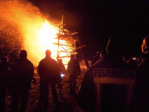 Eine Gruppe von Menschen beobachtet ein großes Feuer, das in der Nacht brennt. Im Hintergrund ist eine Holzstruktur zu sehen, die in Flammen steht.