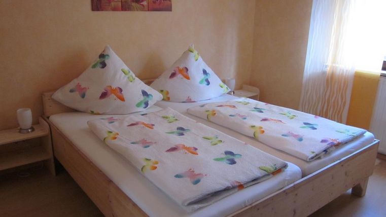 Een gezellige slaapkamer met een tweepersoonsbed en kleurrijke kussens. De muren zijn geel geschilderd en er is helder daglicht dat door de gordijnen stroomt.