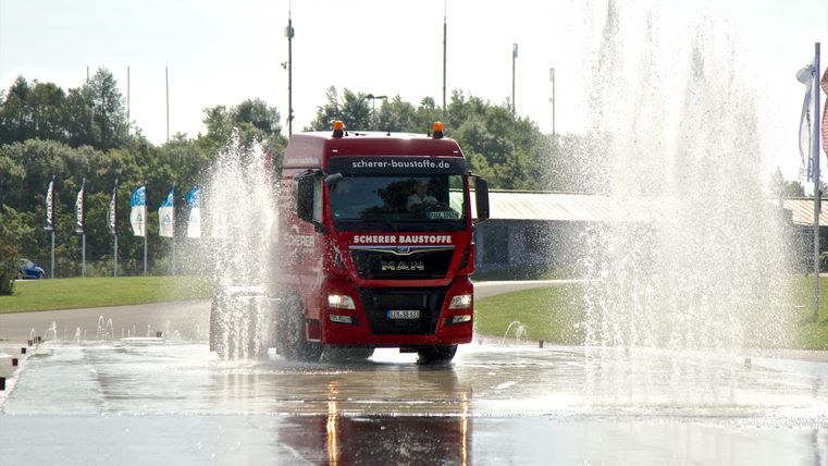Ein roter Lkw fährt durch Wasserfontänen. Die Szenerie wirkt dynamisch und erfrischend.