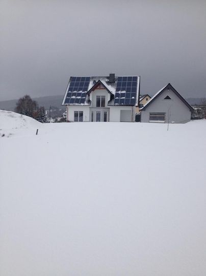 A modern house with solar panels on the roof in a snow-covered landscape. The sky is gray and overcast.