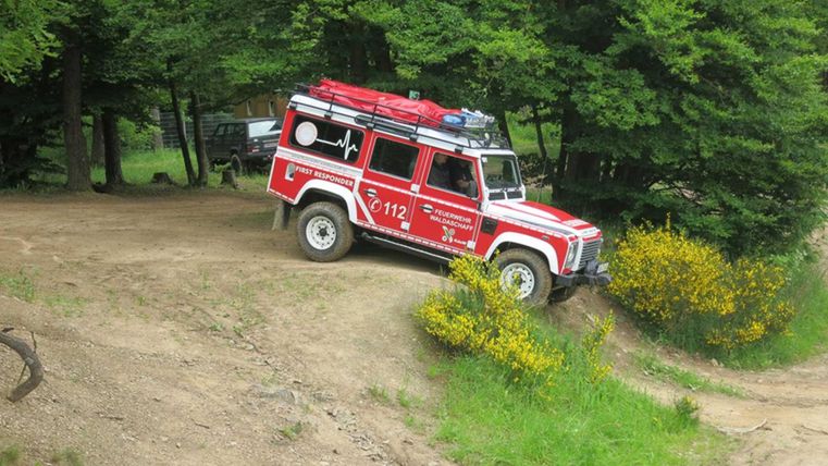Ein rotes Einsatzfahrzeug fährt einen unebenen Weg im Wald entlang. Die Umgebung ist grün und es gibt einige gelbe Blumen.