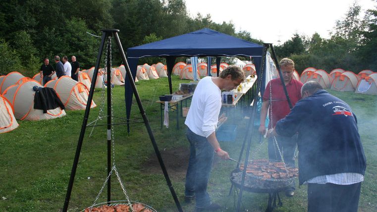 An outdoor barbecue party with several people. In the background, there are tents and green trees visible.