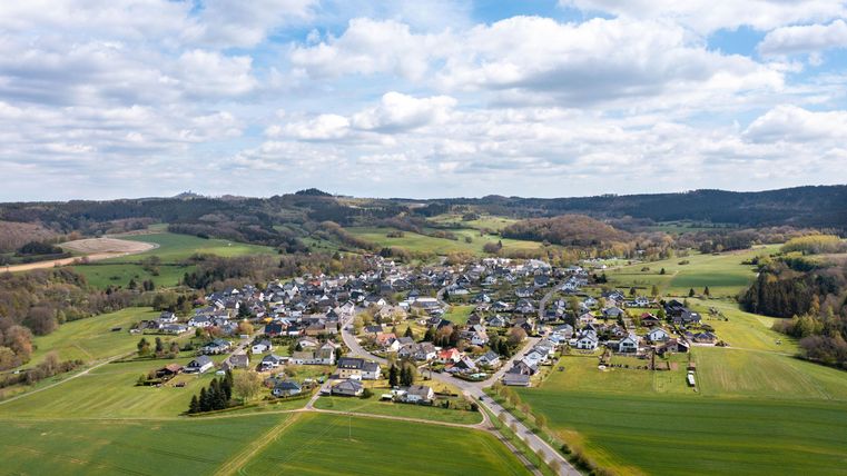 Eine malerische Ansicht eines kleinen Dorfes umgeben von grünen Feldern und Hügeln. Der Himmel ist teilweise bewölkt und der Ort wirkt ruhig und einladend.