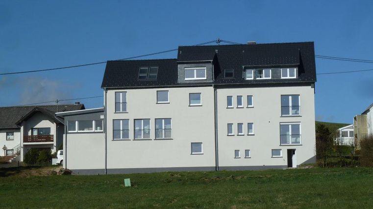 Ein modernes, mehrstöckiges Gebäude mit hellem Anstrich und einem schwarzen Dach. Im Vordergrund befindet sich ein grüner Wiesenbereich.