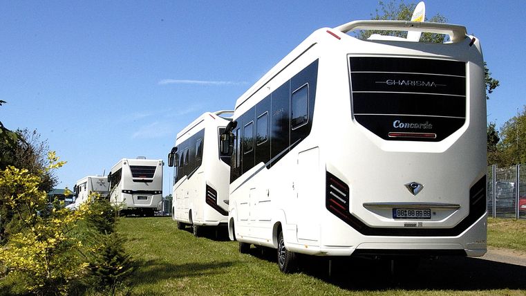 A series of white motorhomes are parked on a grassy area. The sky is clear and blue.