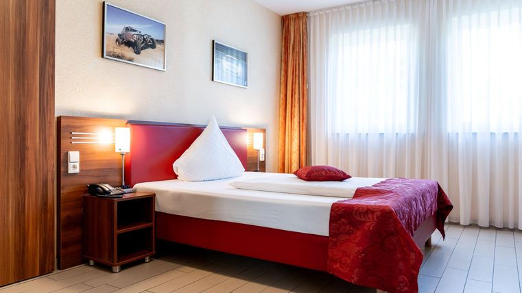 Een gezellig hotelkamer met een tweepersoonsbed en rode beddengoed. Heldere ramen met gordijnen zorgen voor een vriendelijke sfeer.