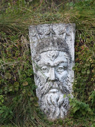 Ein Steinrelief eines bärtigen Mannes mit einer Krone, das von Gras umgeben ist. Die detailreiche Darstellung zeigt eine eindrucksvolle Gesichtszüge.