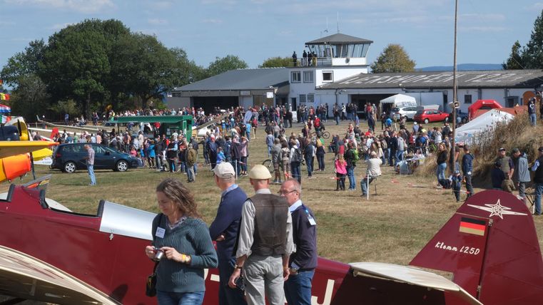 Eine belebte Flugplatzveranstaltung mit vielen Menschen und verschiedenen Flugzeugen. Im Hintergrund sieht man das Hauptgebäude und einen Kontrollturm.