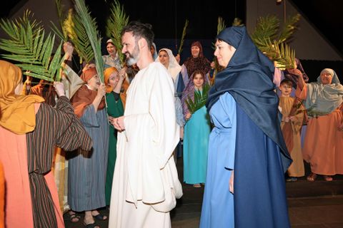 Eine Gruppe von Menschen in historischen Kostümen hält Palmzweige. Im Vordergrund steht eine leicht bekleidete Person, während andere im Hintergrund jubeln.