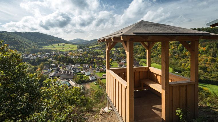 Ein Holzwohnraum mit Aussicht auf das Tal und die umliegenden Hügel. Im Hintergrund ist ein kleines Dorf in einer grünen Landschaft zu sehen.