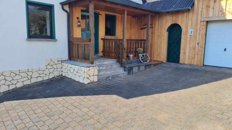 Een uitnodigende ingang van een huis met houten bekleding en een betegelde binnenplaats. Naast de veranda staat een mooi houten wiel.