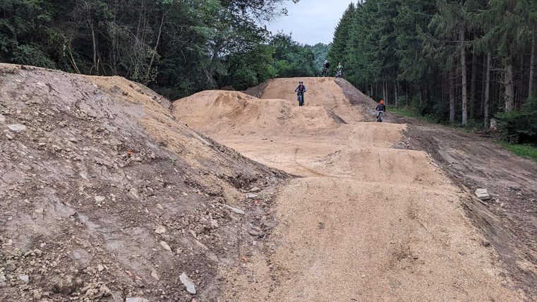 Eine Mountainbike-Strecke mit Hügeln aus Erde im Wald. Zwei Radfahrer fahren auf der Strecke.