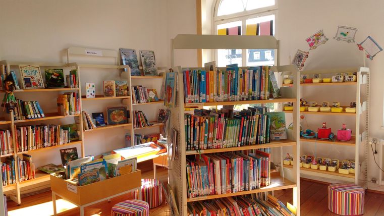 Ein gemütlicher Raum mit Regalen voller Bücher für Kinder. Viele bunte Sitzkissen und dekorative Elemente schaffen eine einladende Atmosphäre.