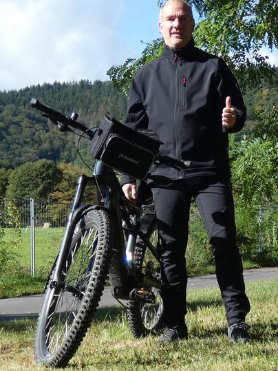 Ein Mann steht neben seinem Mountainbike und gibt einen Daumen nach oben. Im Hintergrund sind Bäume und eine grüne Landschaft zu sehen.