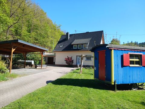Une belle maison avec un carport et une roulotte colorée dans le jardin. Entourée d'arbres verts et d'un ciel clair.