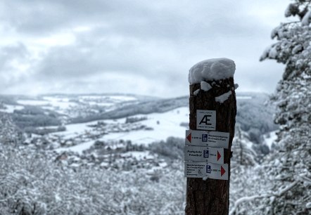 Ein verschneiter Wegweiser im Vordergrund zeigt Wanderwege an. Im Hintergrund erstreckt sich eine winterliche Landschaft mit schneebedeckten H&uuml;geln., &copy; TI Hocheifel-N&uuml;rburgring,S.Schulte