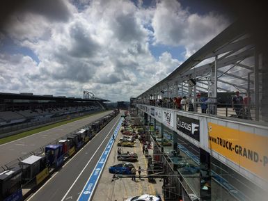 Eine Rennstrecke mit Boxengasse und mehreren Rennwagen. Der Himmel ist teils bewölkt mit einigen blauen Stellen.