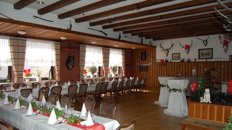 Ein festlich dekorierter Veranstaltungsraum mit langen Tischen und Stühlen. Die Wände sind mit Geweihen und Bildern geschmückt, und es gibt große Fenster mit Vorhängen.