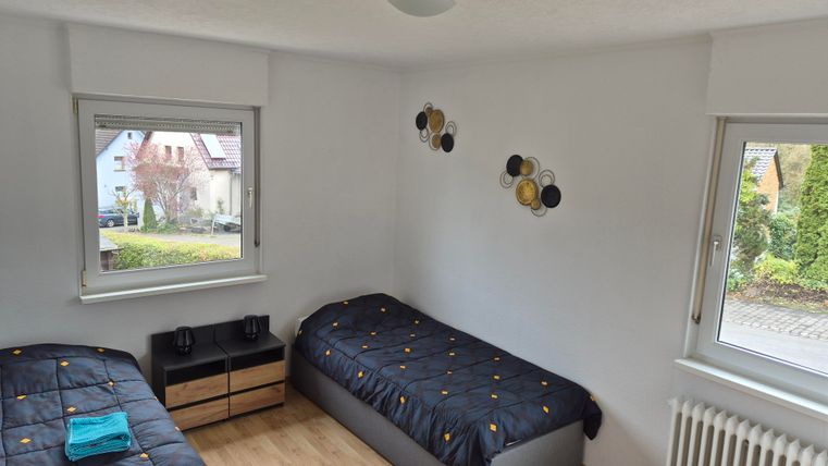A bright bedroom with two single beds and a simple nightstand. Large windows offer a view outside and provide plenty of light.