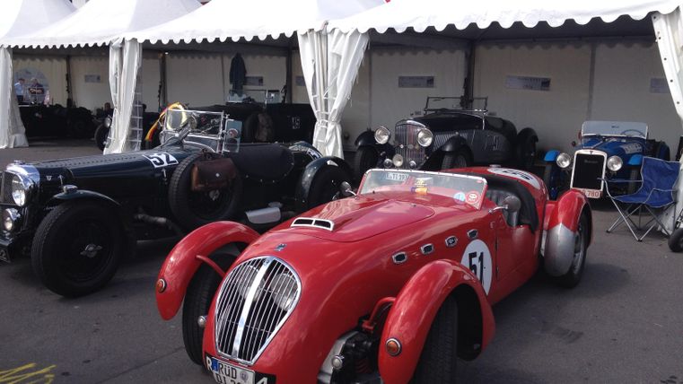 Ein rotes Oldtimer-Auto parkt vor einem Zelt. Im Hintergrund sind weitere klassische Fahrzeuge zu sehen.