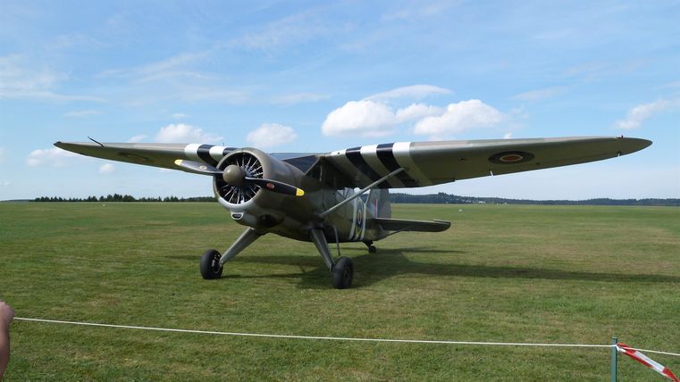 Ein altes Flugzeug steht auf einem grünen Feld. Der Himmel ist blau mit wenigen Wolken.