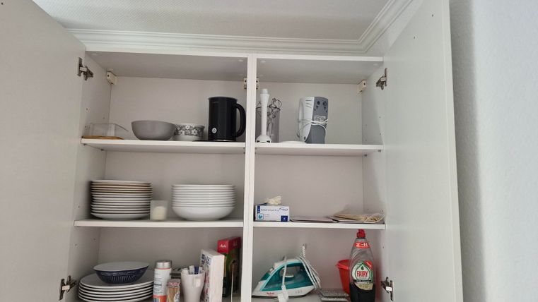 A kitchen cabinet with several shelves containing dishes, cups, and kitchen appliances. Some items like a coffee maker and an iron are also visible.