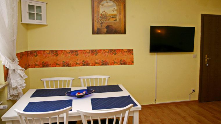 Een gezellige eetruimte met een witte tafel en vier stoelen. De muren zijn geel geschilderd en een televisie en een schilderij zijn aan de muur gemonteerd.