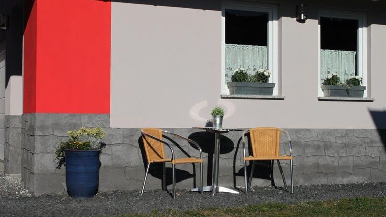 A small table and chair set is in front of a house with red and grey color accents. The windows are decorated with flower boxes.
