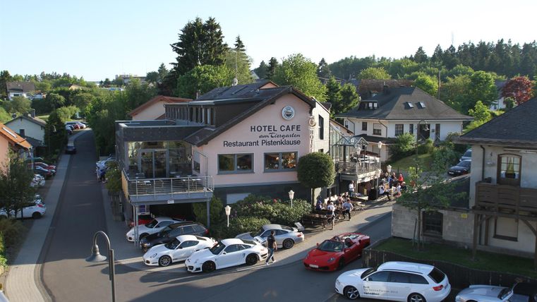 A picturesque hotel with a café and restaurant in a green setting. Several cars are parked nearby, and the outdoor area is well frequented.