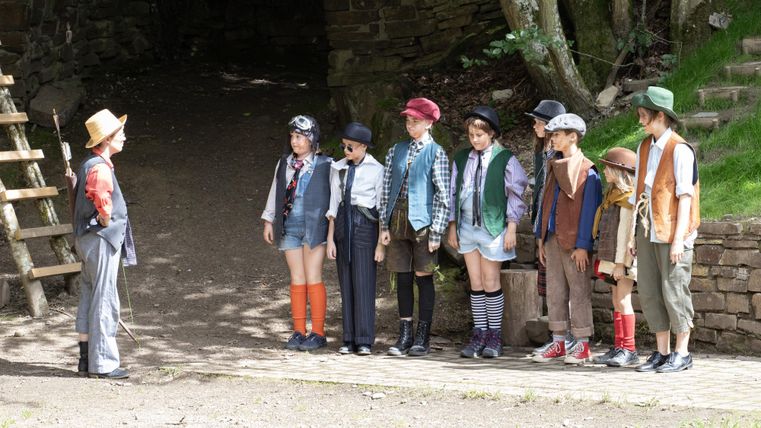 Eine Gruppe von Kindern steht in einer Reihe im Freien, während ein Kind mit einem Hut ihnen gegenübersteht. Sie tragen verschiedene Kostüme und scheinen an einer Theateraufführung teilzunehmen.