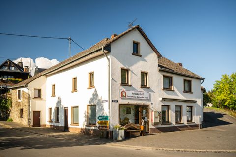 Een charmant, wit gebouw aan een straat. Het is een herberg met een uitnodigende ingang en omgeven door bomen.