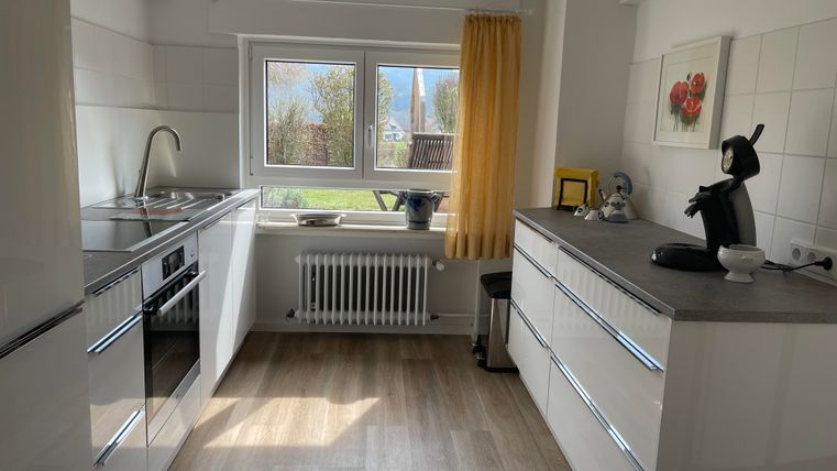 Une cuisine moderne avec des armoires blanches et un sol en bois. Il y a des fenêtres avec vue sur l'extérieur et quelques appareils de cuisine sur le plan de travail.