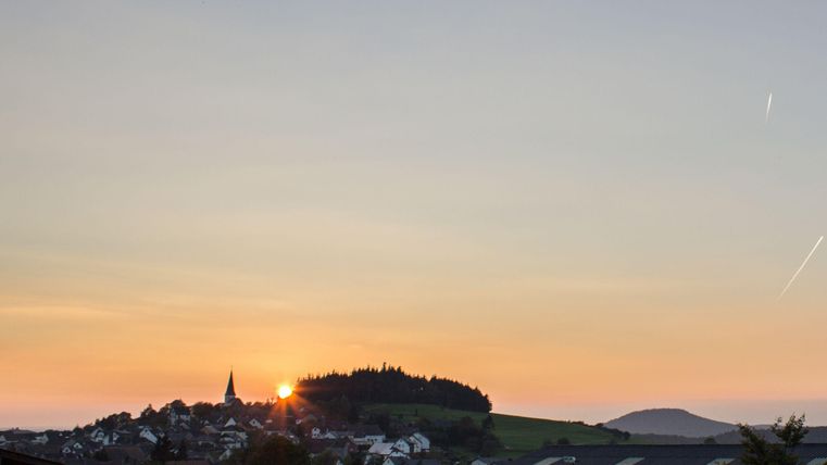 Un paysage pittoresque avec un coucher de soleil derrière une colline. Au premier plan, on peut voir des maisons et des arbres.